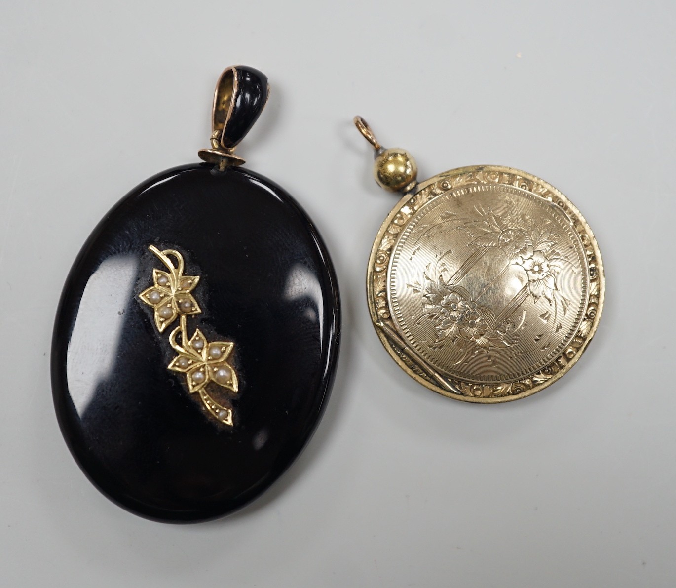 A Victorian yellow metal and seed pearl mounted black onyx oval pendant locket, with enamelled bale, overall 53mm and an engraved gold plated circular pendant locket.